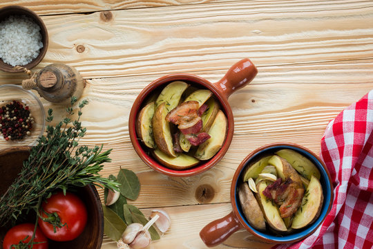 fried potatoes and bacon in ceramic forms on wooden table with clay plate with tomatoes,  thyme, rosemary, sea salt, olive oil in clay jug, garlic,  pepper, bay leaf , red squared tablecloth, top view