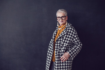 Stylish old woman in glasses standing on a black background.