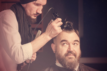 Hairdresser in a black cap and a black apron cuts with hairclipper a man with a clip on the head