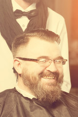 Stylist in black scarf and bow tie makes hair a man with a beard and glasses