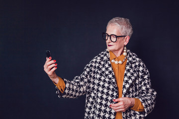 Stylish and elegant elderly woman in glasses makes selfie on a black background.