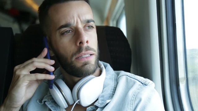 Giovane ragazzo con la barba sta parlando al telefono seduto sulla poltrona di un treno con le cuffie appese al collo