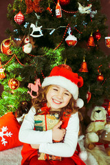 Little girl in red dress sits with a present box before a Christmas tree