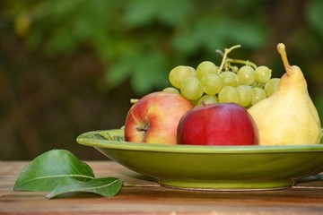 Apples, grapes and pear in a bowl on a background of green leaves.