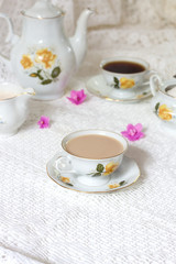 Obraz na płótnie Canvas A tea cup decorated with flowers, two biscuits and a candle on a white wooden table. In the background a turquoise clock on a yellow wainscot. Vintage tea time