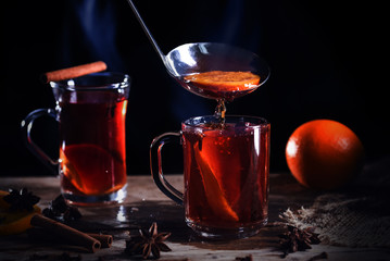 hot steaming mulled wine is poured into glass mug, with Christmas spices like orange slices, cloves, star anise and cinnamon on a rustic wooden table against dark background