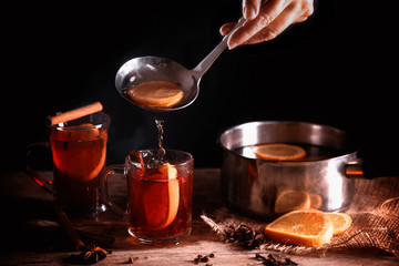 pouring hot mulled wine from in a pot and in glass mugs, Christmas spices like orange slices, cloves, star anise and cinnamon on a rustic wooden table against a dark background