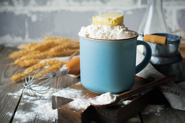 Ingredients and tools for homemade baking, flour and butter in a ceramic cup on an old wooden background. Selective focus.