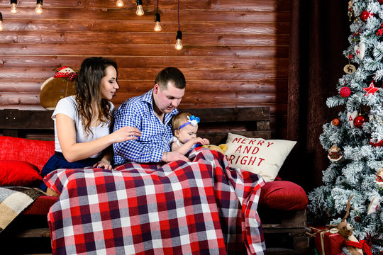 Portrait Of Happy Family With Little Beautiful Daughter Sitting On The Wooden Sofa With Warm Red Blanket Against Wooden Wall, Near The Christmas Tree. Concept Of Celebration And New Year