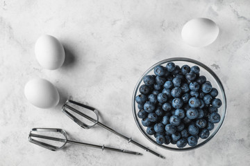Top view photo of blueberries in a glass plate. Mixer fits and eggs on a white surface. Ingredients for healthy rustic breakfast.