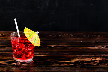 Top view of glass of red cocktail with ice cubes and straw umbrella on dark wooden table, close-up, copyspace for text