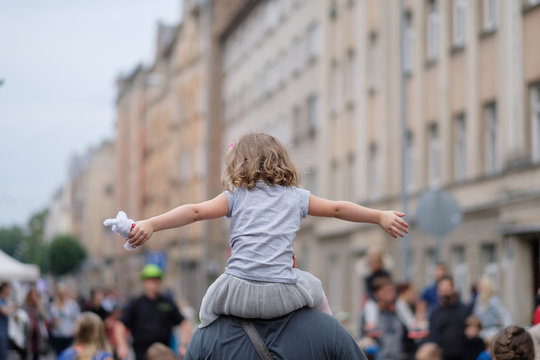 A girl with arms spread sits on her father's shoulders at a city party