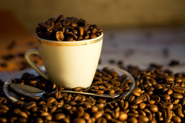 white coffee cup laid on a wooden table sprinkled with coffee beans in darkness illuminated by a side light