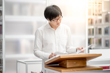 Young Asian man university student reading recommended book on podium in library, education...