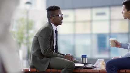Tracking of African businessman sitting on bench outdoors and chatting with male Asian colleague during break; young man eating sandwich and drinking coffee - Powered by Adobe