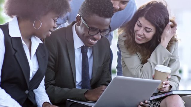 Tilt up slowmo shot of diverse group of colleagues watching something on laptop and laughing while sitting together outdoors
