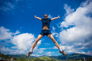 Alone man Jumping for Joy on a Grass Hill above horizon line. Happy slim man fly in a green field against blue summer sky with clouds and mountains. Idea of success, growth, light, diet, luck, freedom