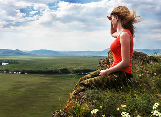 The girl sits on the mountain and looks into the distance. At the bottom are green fields.