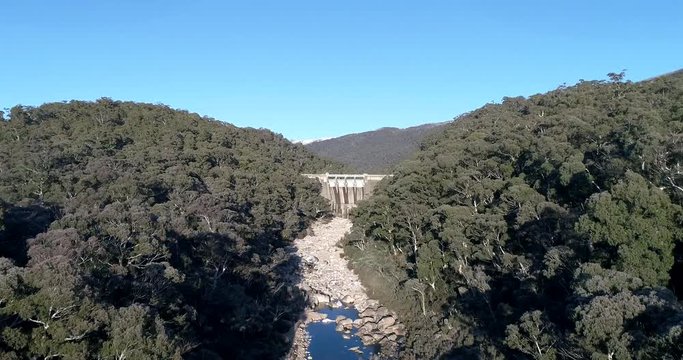 Snowy river from riberbed to Hydro dumb between evergreen gumtrees under blue sky.