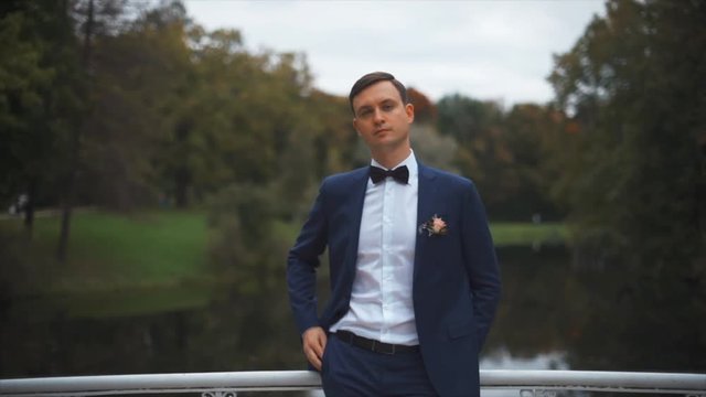 Close up portrait of handsome stylish groom outdoors in park with dark red bowtie