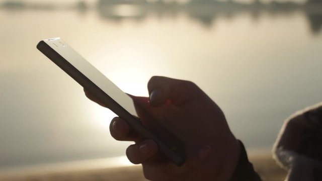 Sunset, On the Background of the Sea A beautiful sexy girl sits on the beach near the sea, wrapped in a blanket, uses a smartphone, social networks and crypto currency - bitcoin.