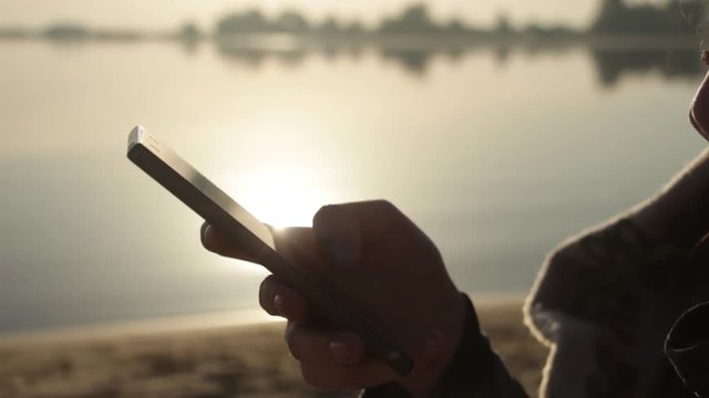 Sunset, On the Background of the Sea A beautiful sexy girl sits on the beach near the sea, wrapped in a blanket, uses a smartphone, social networks and crypto currency - bitcoin.