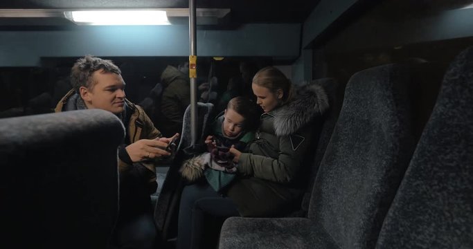 Family of three traveling by bus in winter evening. Man making photo of his wife and son playing on cell during the ride