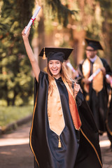 The young happy joyful graduate in mantle standing outdoor and keeping diploma in hand and smiling