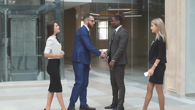 Confident and attractive business team of mixed ages and ethnicity meet and shake hands in the lobby of a busy modern office building.
