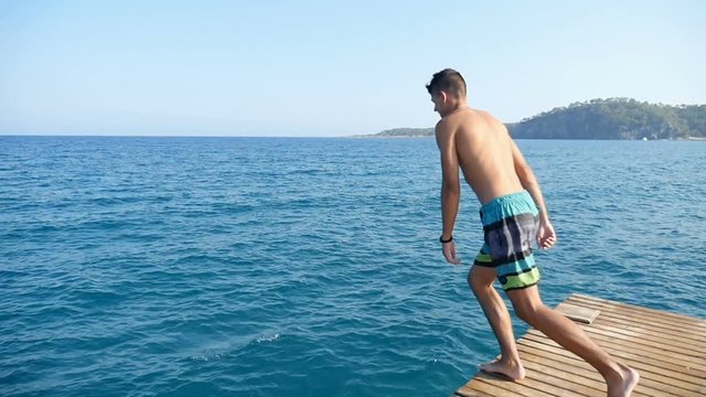 A sportive young man in multicolored shots jumps in the Mediterranean sea waters feet first in slow motion. The seascape with sparkling blue waters looks great and gorgeous
