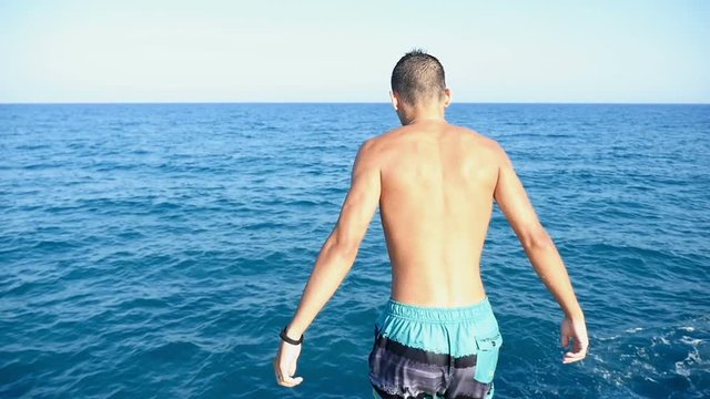 An athletic young man in multicolored shots jumps in the Mediterranean sea waters feet first in slow motion. The seascape with sparkling blue waters looks great and gorgeous