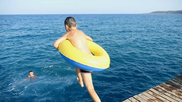 A seven-year-old boy with a yellow and blue inflatable ring jumps into the Mediterranean sea from a pier in summer in slow motion. The horizon line looks fine