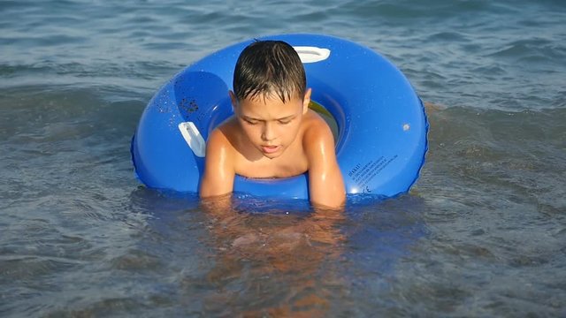 A seven-year-old boy swims in a blue inflatable ring in the dark waters of the Mediterranean sea in summer in slow motion. He looks engaged and entertaining