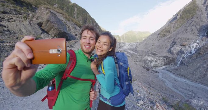 Selfie couple taking phone self-portrait on New Zealand by Franz Josef Glacier. New Zealand tourists smiling happy in nature by Franz Josef Glacier, Westland Tai Poutini National Park.