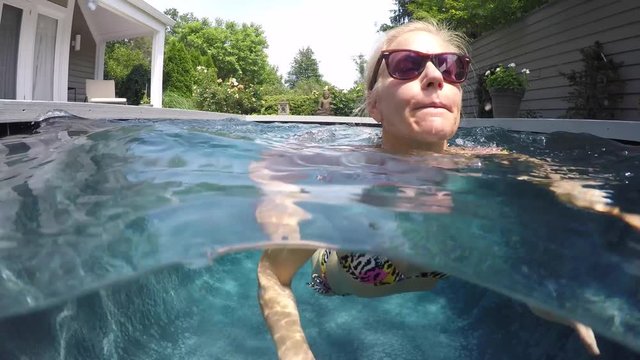 Camera half above and half below water, a beautiful, slim, caucasian woman wearing string bikini swims toward the camera.