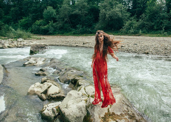 A sweet girl stands on a rock in the middle of a boiling river. The wind twists her long hair beautifully. Background wild nature. Creative colors.