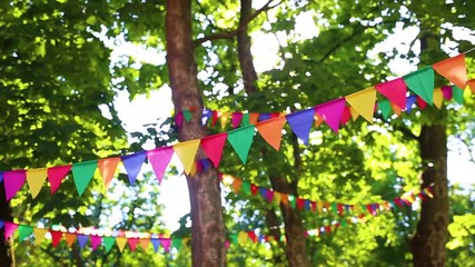 Colorful flags hanging on branches of trees as festive decoration for outdoor party. Bright summer sunshine among green leaves in background. Real time full hd video footage.