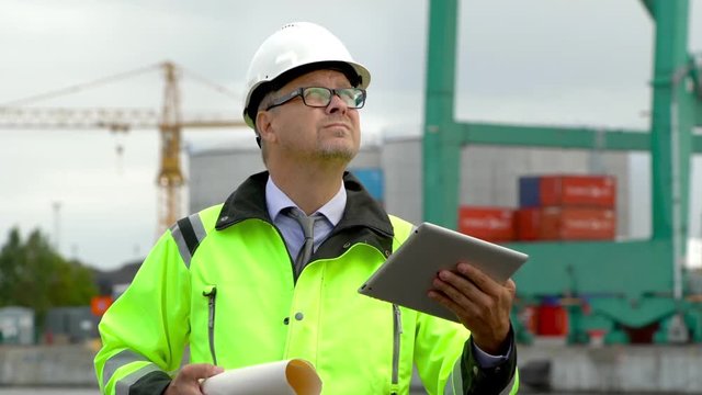 A foreman planning logistics, using a tablet and holding a drawing, in front of a container terminal