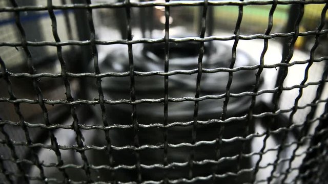 Coil with metal wire rotates behind grate on machine. At factory machine works from which rods come out for production of product.