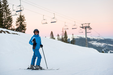 Fototapeta na wymiar Happy skier female in blue jacket, ski pants and goggles on her head standing in the snowy mountains enjoying winter holidays against a ski-lift