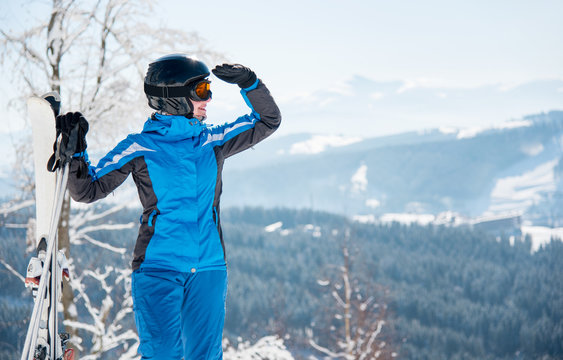 Close-up portrait of female skier with skis enjoying stunning scenery in the mountains looking away, wearing blue ski suit and black helmet copyspace
