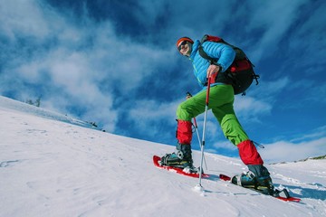 Climber with backpack trekking poles snowshoes rises to the top of the mountain in the snow on a background of beautiful blue sky.
