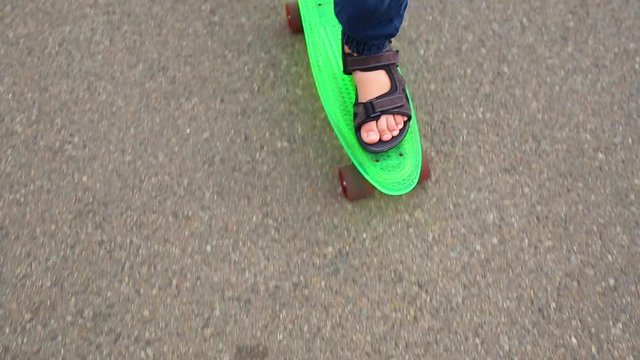 Point of view shot of male feet of child skating on green plastic penny-board. White kid of 10 years has fun in city park on warm summer or autumn day. Top view real time full hd video footage.