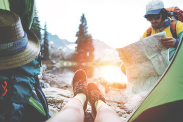 Young couple camping together in stunning mountain wilderness near the lake. POV from the tent. Woman sitting inside, man wearing sunglasses and backpack and searching for trails on the map