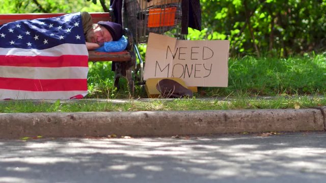 Tramp lying on bench using USA flag as blanket. Homeless aold male sleeping on bench, board with sign need money on ground close to bench