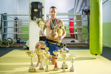 Young boxer champion with many medals around his neck posing below his trophy cups in sports hall.