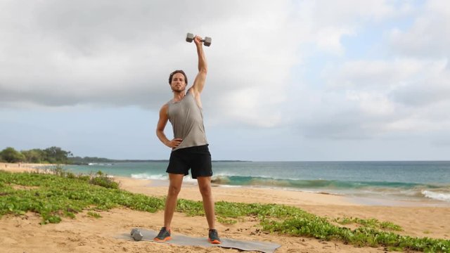 Fitness Dumbbell Snatch - man doing strength training exercises with Dumbbells on beach. Fit handsome male fitness model sweating during dumbbell workout exercise outdoors on beach. SLOW MOTION.