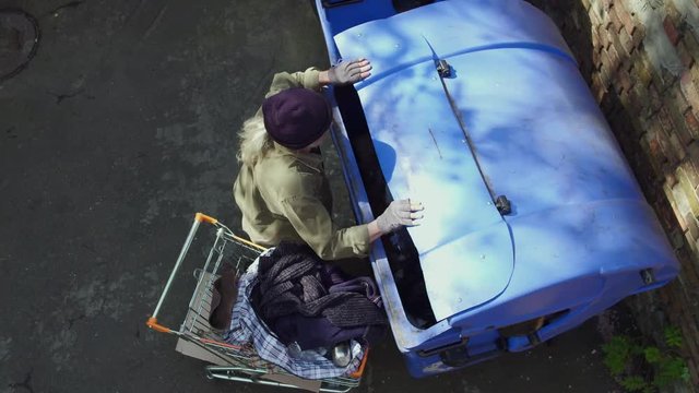 Top view of homeless man approaching trash can with shopping cart. Tramp digging in garbage in search for food and empty bottles.
