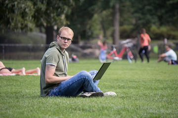 Fototapeta premium Young man in glasses working on laptop sitting on the grass in park. Maybe student or hacker