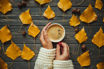 Woman on knitted sweater holding hands cup cappuccino on  wooden table. Autumn leaves and acorn.Top...